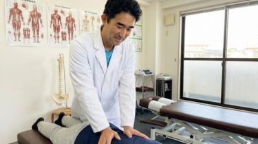 白衣を着た男性カイロプラクターが、診療室で患者の腰部に手技療法を行っている笑顔の様子。背景には人体の解剖図と脊柱模型が見える。