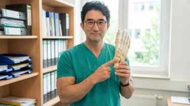 緑色のスクラブを着て眼鏡をかけたアジア人男性医師が、オフィスで足の骨格模型を持ち、左手の人差し指で足首付近の骨を指差している写真。