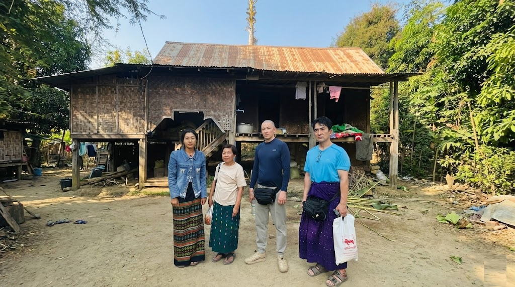 ミャンマーのタウンドィンジー村にて、カイロプラクティックのボランティア治療活動に参加した日本人男性2人と、現地の女性2人が、伝統的な竹造りの高床式住居の前に並んで立っている様子。