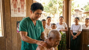 緑のスクラブを着た筋肉質の日本人男性（榊原学長）が、ミャンマーの現地の診療所で高齢男性に徒手療法を行っているボランティア活動の様子。背景には解剖図が貼られ、現地の女性や子供たちが笑顔で見守っている。