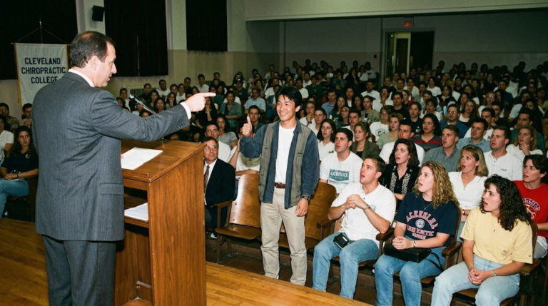 カイロプラクティック大学（アメリカ）のオリエンテーション講堂で、壇上の学長に見事名前を言い当てられた日本人留学生が笑顔でサムズアップをし、周囲のクラスメイトたちが驚いてどよめいている様子
